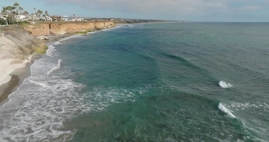 An aerial shot of a coastline with houses on the cliffs and the sea with rolling waves and a cloudy sky