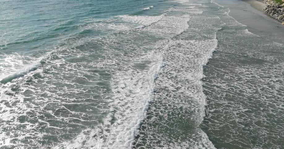 Sunlit waves create rippling white foam patterns as they roll toward the sandy shore
