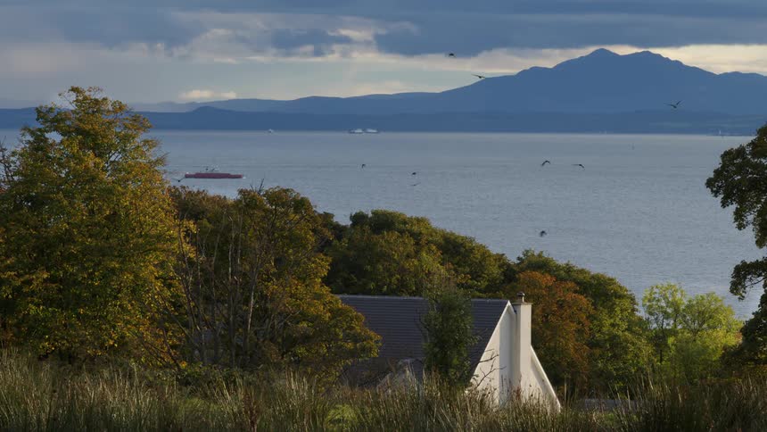 A serene landscape view showcases a white house nestled among autumn trees overlooking a vast body of water with distant mountains ships and birds flying under a cloudy sky