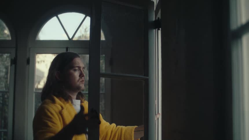 Man in yellow robe closing a window in an indoor space lit by natural light