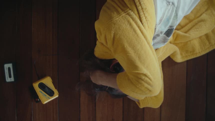 A person lying on a wooden floor with their head propped up by their hands listening to a yellow walkman while wrapped in a yellow robe