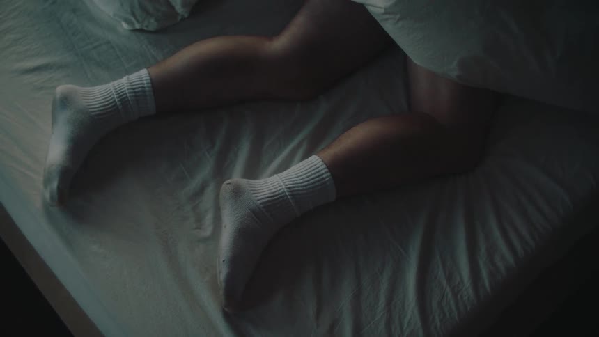 A persons legs wearing white socks as they lie and shift slightly on wrinkled white bedsheets
