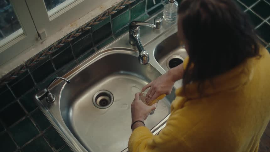 A person in a yellow robe pouring orange juice from a glass into a doublebasin kitchen sink then rinsing the glass with water
