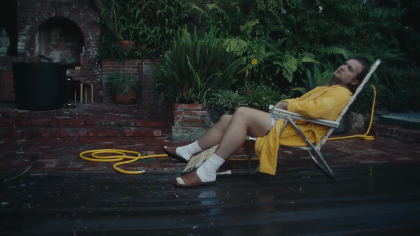 A man in a yellow robe lounges in a folding chair outside on a wet patio near greenery
