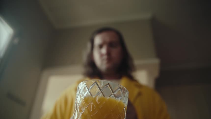 A lowangle shot shows a man in a yellow robe pouring orange juice from a carton into a glass the camera focuses on the liquid filling the glass which has a textured faceted surface
