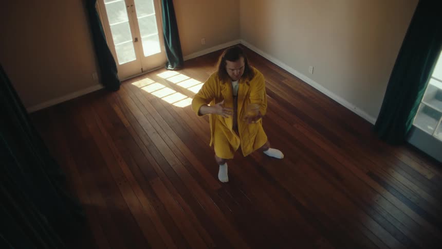 A longhaired man in a yellow robe performs a series of choreographed movements reminiscent of tai chi or a martial art the man moves with a deliberate and flowing motion with a sunlit wooden floor and white walls as the backdrop