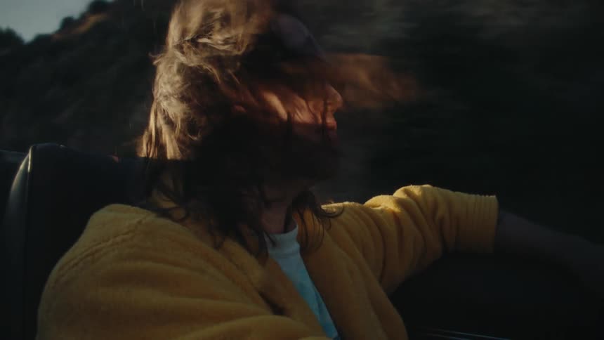 Hair blows around while a man drives his convertiblepast a hill with the top down