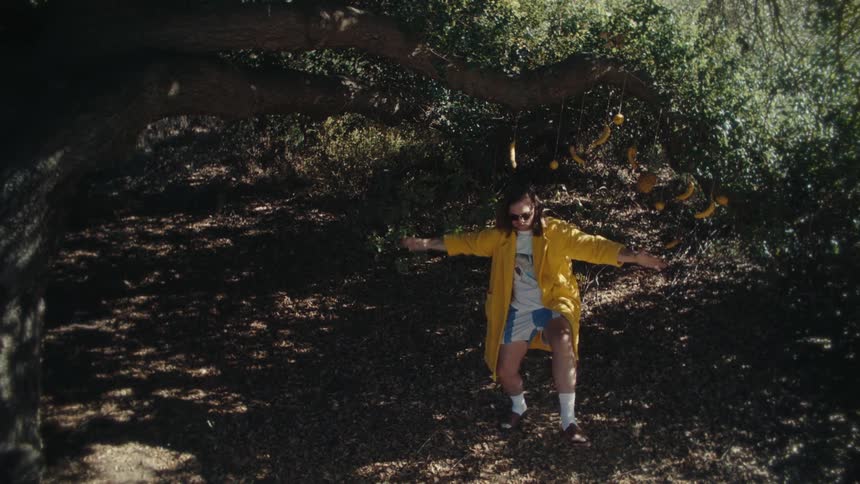 A young man wearing a yellow jacket and sunglasses dances excitedly in the woods at midday
