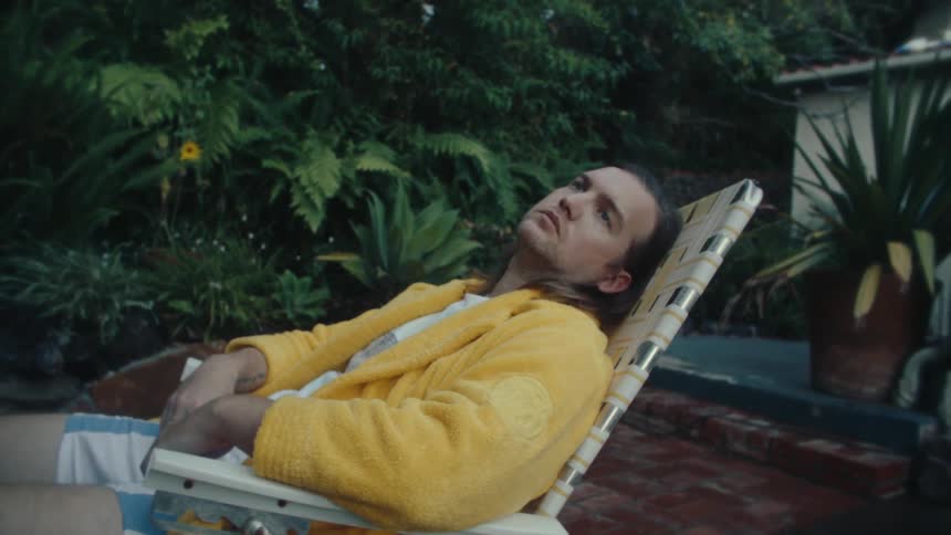A man wearing a yellow jacket sits on a pool chair in the backyard of a building surrounded by trees deep in thought and looking sad
