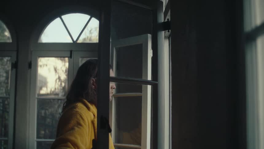 Person in a yellow robe opening a window in an indoor setting lit by outside light