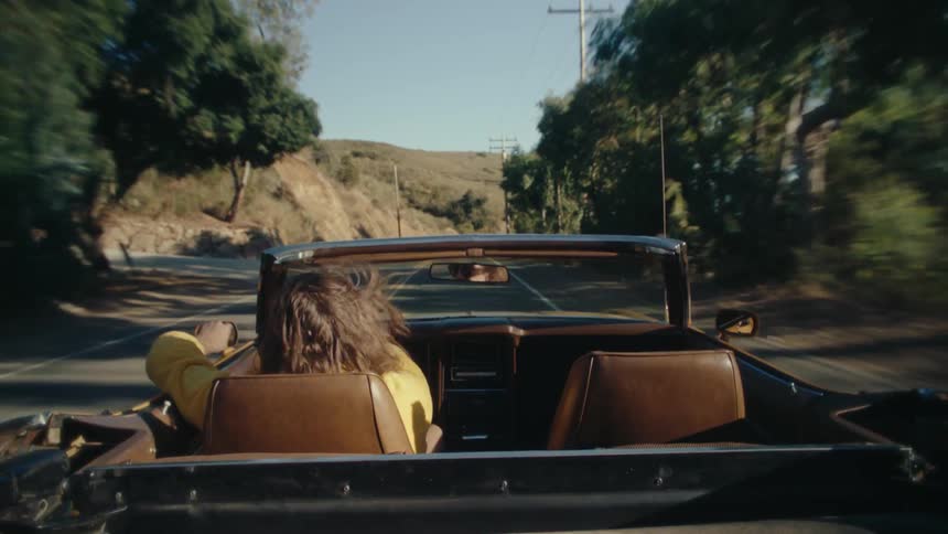 The video shows a person with long brown hair wearing a yellow robe riding in the passenger seat of a convertible car as the car moves along a winding road the persons hair blows in the wind