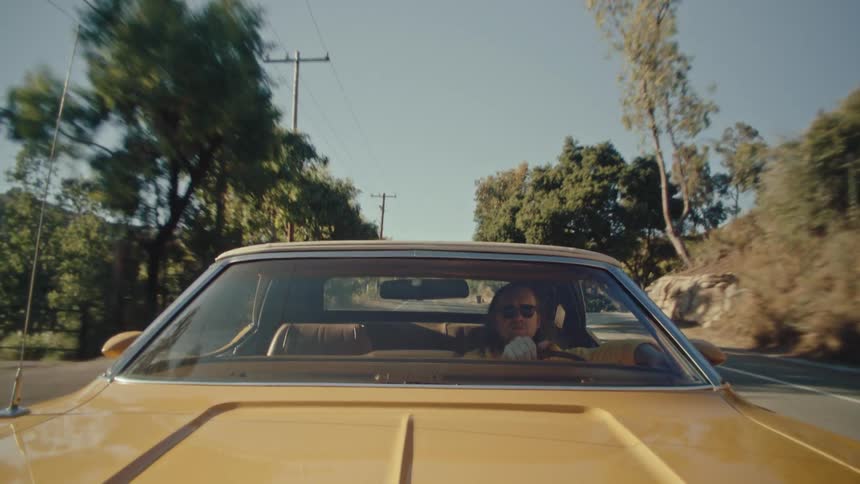 A wideangle shot from the front of a classic yellow car showing a person with long hair driving down a sunny treelined road