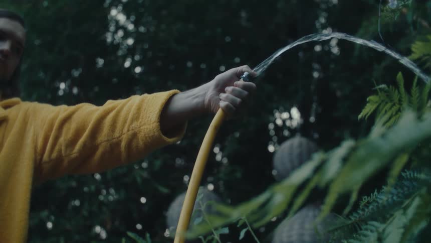 A person wearing a longsleeved yellow shirt uses a water hose to water plants in a garden the hose has a nozzle that creates a wide arching spray of water which is directed towards some lush green leaves