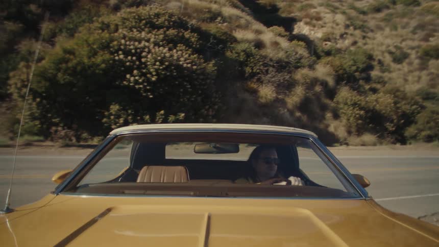 The video shows a lowangle shot from the perspective of a person in the passenger seat of a car the camera is aimed toward the front of the car with the roof and side of the car visible as the car drives through a hilly arid landscape