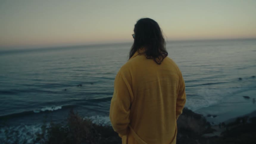 A person with long hair wearing a yellow robe standing on a cliffside overlooking the ocean at dusk