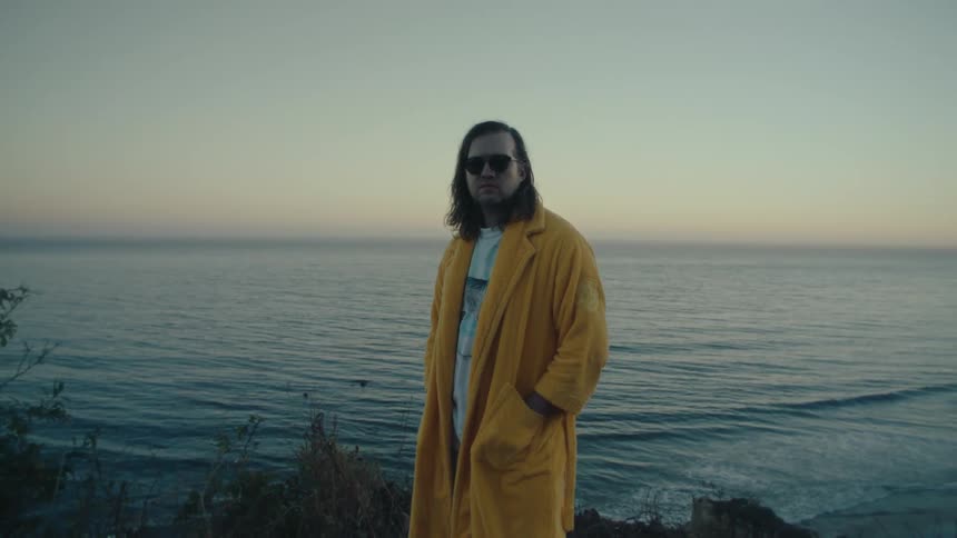 A person in a yellow coat stands on a coastal cliff at sunset