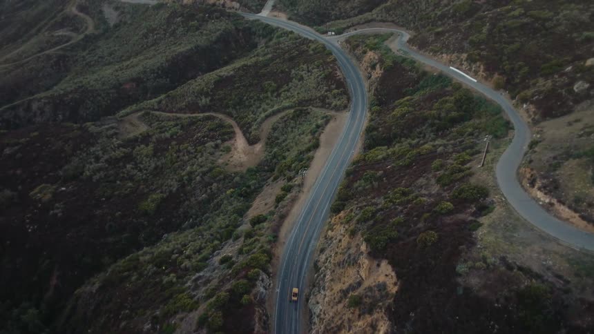 An aerial drone shot captures a yellow car driving along a winding road that snakes through a hilly arid landscape the camera remains stationary allowing the car to drive out of the shot