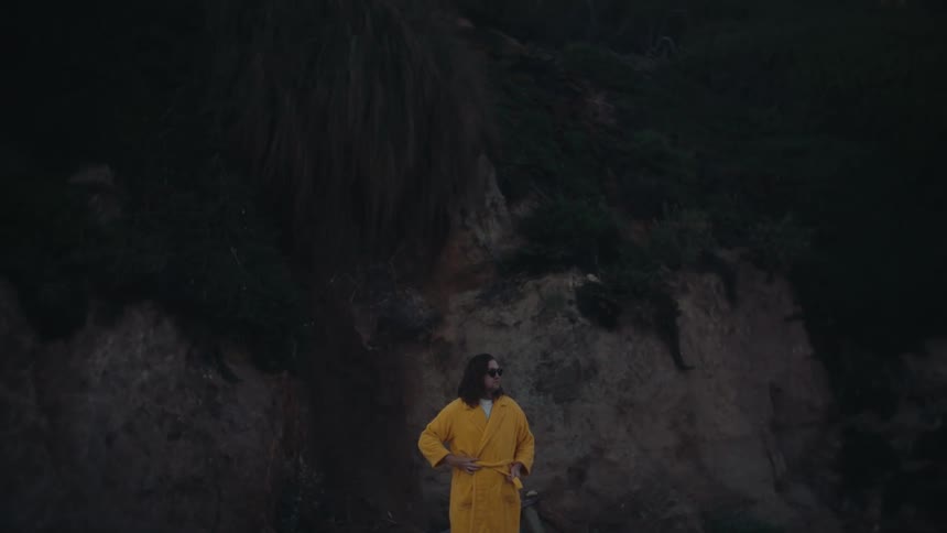 Man wearing a bathrobe stands on the beach near the base of a hill after dusk