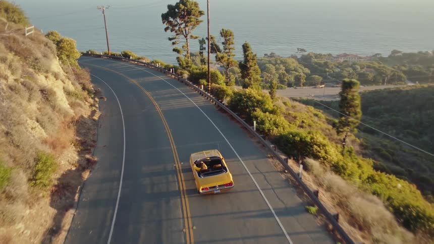 An aerial shot follows a yellow convertible driving down a winding coastal road at sunset