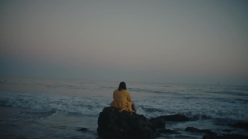 Person wearing a yellow robe sits on a rock formation on the beach while staring towards the red tinted horizon