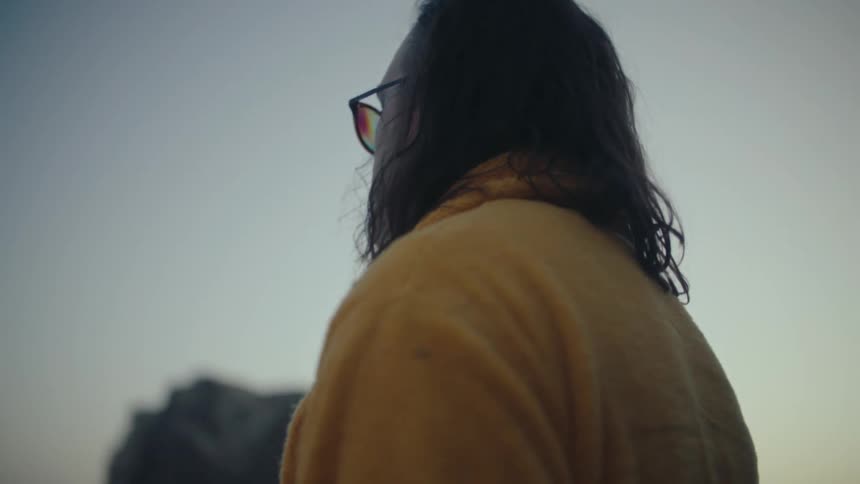 The video shows a man with long dark hair and sunglasses wearing a yellow robe standing with his back to the camera he is looking out at a hilly landscape and the camera slowly zooms out to reveal the surrounding scenery