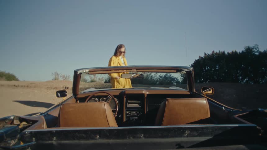A drone shot captures a yellow convertible car driving on a scenic road that winds through a hilly arid landscape the camera then pans and follows the car from a different angle as it continues along the road