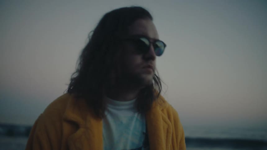 A man in a yellow coat and sunglasses stands by the shore at dusk