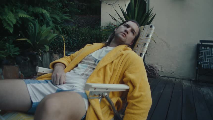 The video shows a man with long hair wearing a yellow robe and white socks relaxing in a lounge chair on a deck a yellow garden hose is visible on the ground next to him
