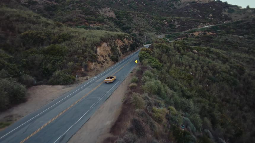 A yellow car driving on a highway in a mountainous region at midday with the roads lined with grass