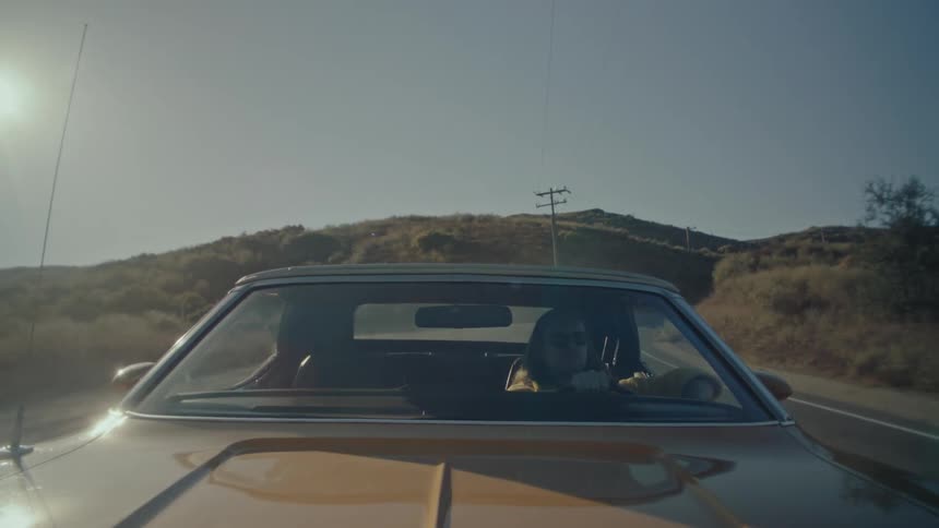 A person drives a vintage convertible down a rural road as the sun flares brightly in the sky