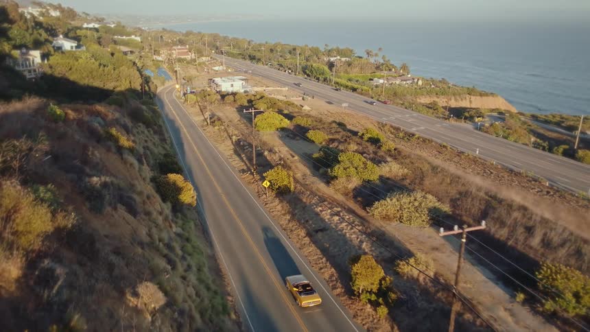 A yellow convertible drives along a scenic road that runs parallel to a larger coastal highway overlooking the ocean