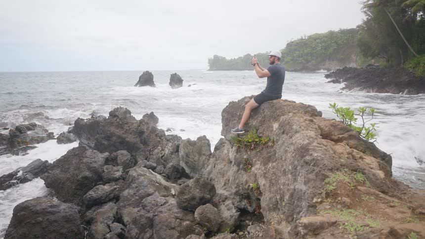 Man sitting on rocks along the coast as he pulls out his phone to take a picture
