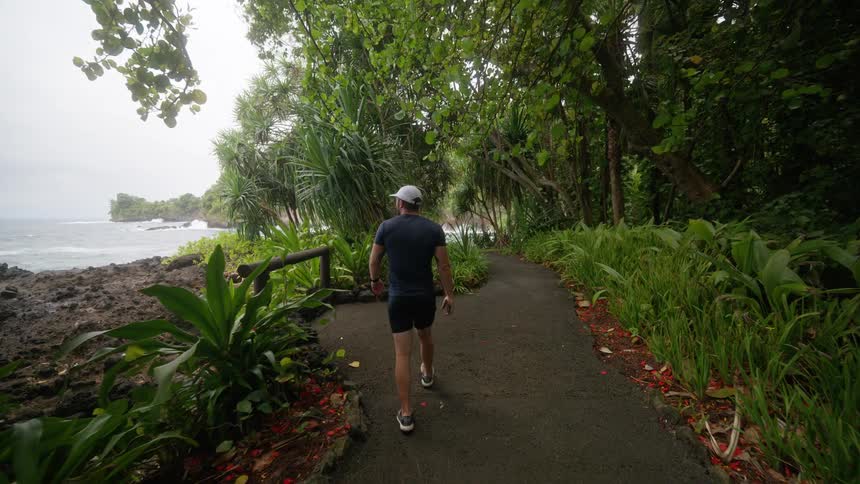 The video shows a person in a dark shirt and shorts walking along a paved path next to a rocky shoreline the path is lined with lush green plants and trees with the ocean visible on the left side of the frame