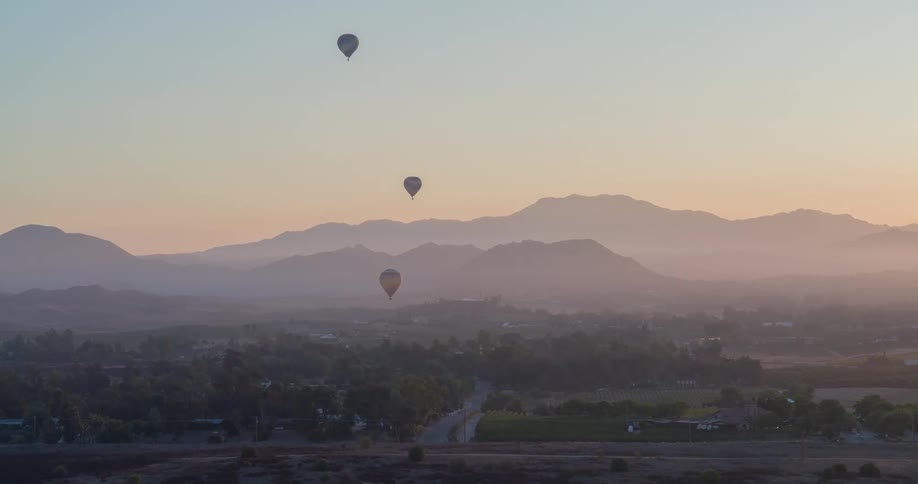Three hot air balloons are floating in a hazy orange and yellow sky during sunrise or sunset over a valley with green trees and a distant mountain range