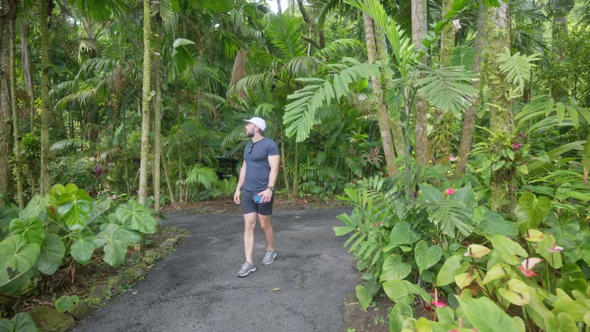 A man wearing a white baseball cap sunglasses a dark blue tshirt and black shorts is walking down a paved path in a lush botanical garden surrounded by tropical plants