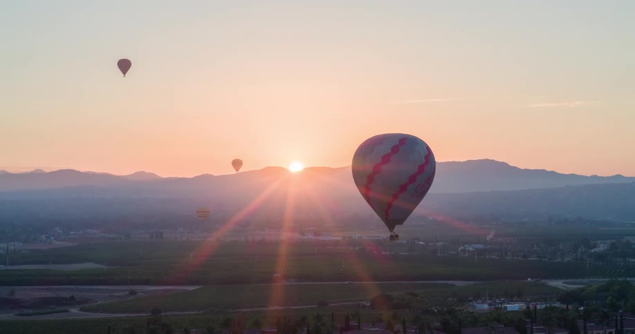The video shows an aerial shot of a hilly landscape at sunrise with two hot air balloons floating in the sky one balloon is striped and the other is a reddishbrown color the sun is visible just above the hills in the background with sunbeams radiatin