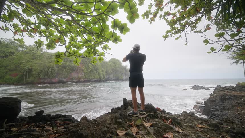 A man stands on a rocky tropical coast taking a picture of the ocean with his phone