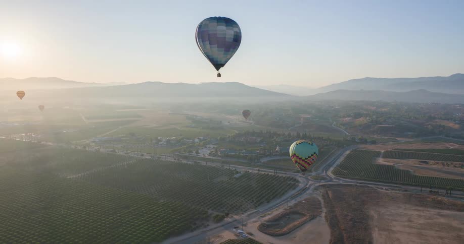 The video shows an aerial shot of a hilly landscape with vineyards and trees two hot air balloons one with a blue and white pattern and another turquoise one are floating above the landscape