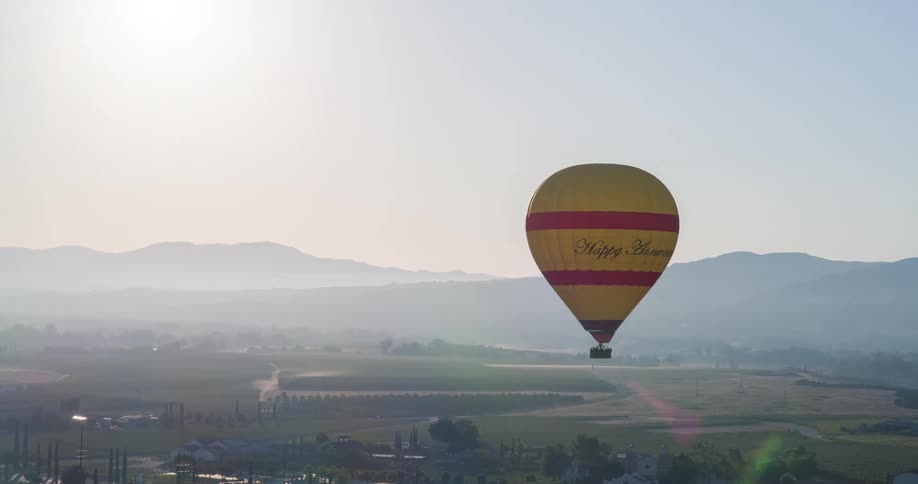 The video shows an aerial shot of a single hot air balloon with yellow and red stripes hovering over a rural landscape the surrounding area consists of vineyards trees and hills shrouded in a hazy morning light