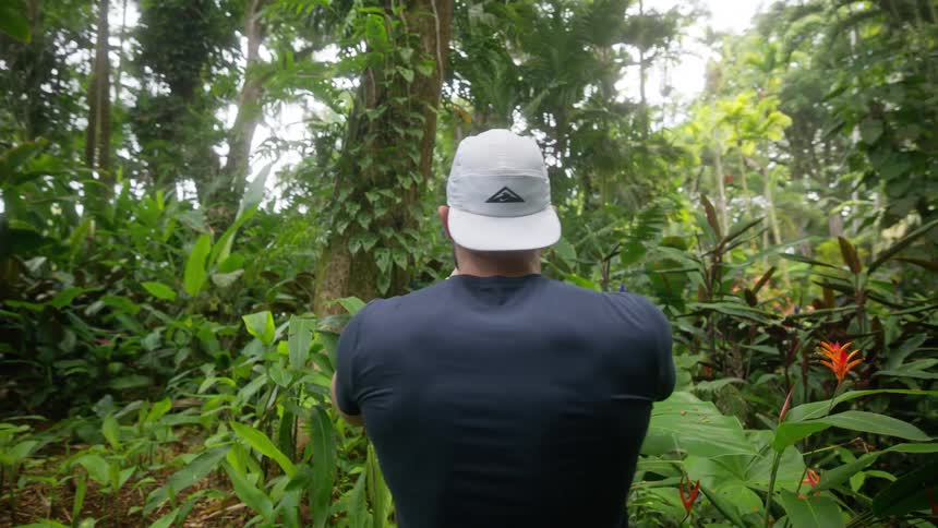 The video shows a man in a dark shirt and shorts with a white baseball cap walking along a paved path in a lush tropical garden the path is surrounded by a variety of green plants and trees