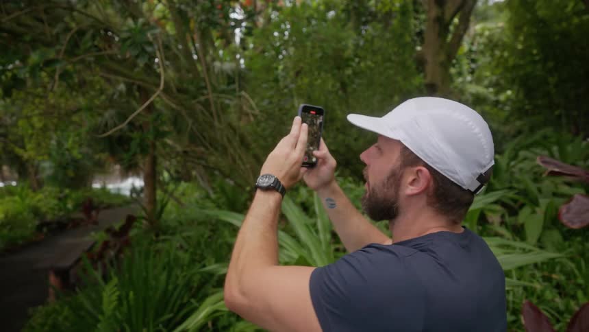 A man wearing a baseball cap walks along a paved path surrounded by trees and lush plants he pauses to take a picture of the ocean with his phone which is visible on the left side of the frame