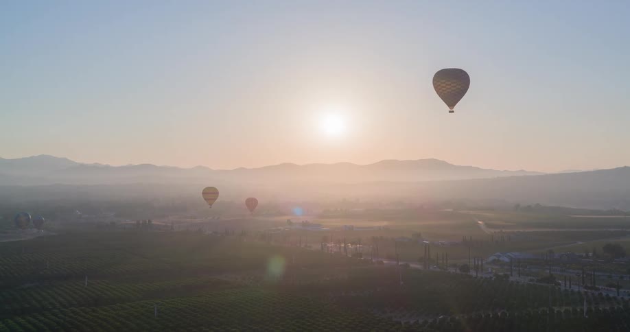 Three hot air balloons are floating in a hazy pale sky as the sun shines illuminating the rolling green hills and distant mountain range below