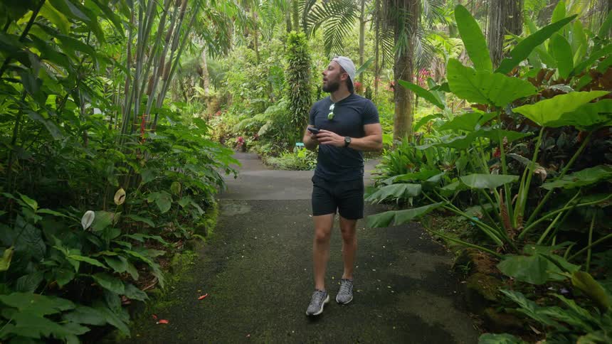 Man wearing a cap and using a smartphone walking through a tropical park