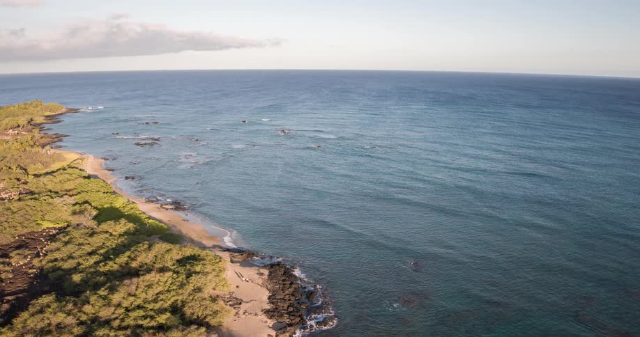 An aerial view shows the bluegreen ocean with breaking waves and a rocky shoreline as the camera slowly moves to reveal a small sandy beach and land covered in green shrubs and dark volcanic rock
