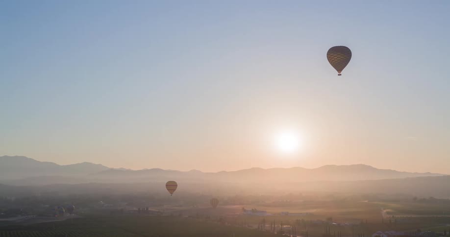 The video shows an aerial shot of a few hot air balloons floating over a vast green landscape during a beautiful sunrise the sun shines brightly over the mountains in the distance illuminating the fields and trees below