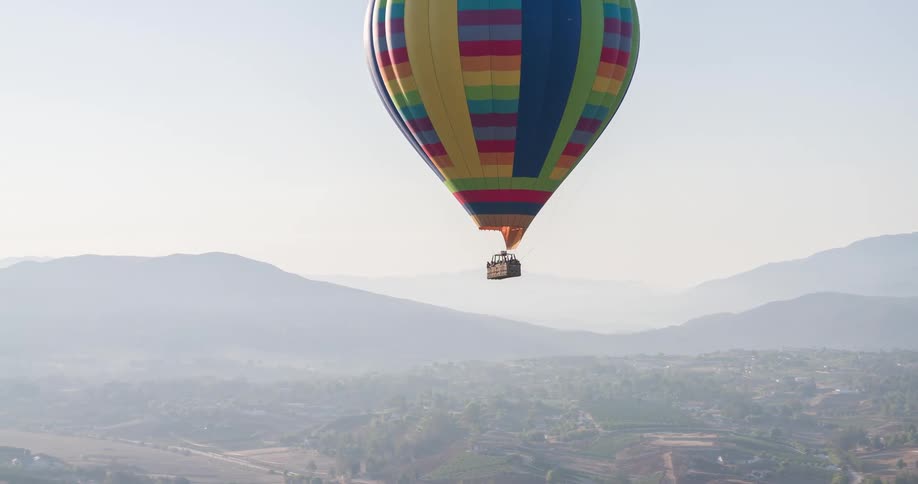 A multicolored hot air balloon is shown from a high vantage point floating in the hazy sky over a green valley with a mountain range in the distance