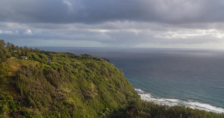 A view from above shows a steep tree covered cliffside next to the ocean with white waves crashing on the shore below a sky filled with gray clouds