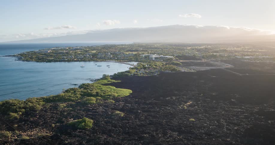 A large bay with dark blue water and several small boats is shown from above with a town green vegetation and black volcanic rock in the foreground and a large mountain range in the distance