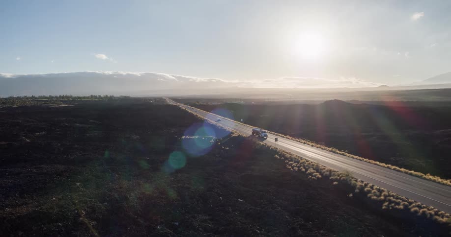 An overhead view of a truck driving on a long straight road stretching through a barren rocky landscape toward a sunlit mountain range