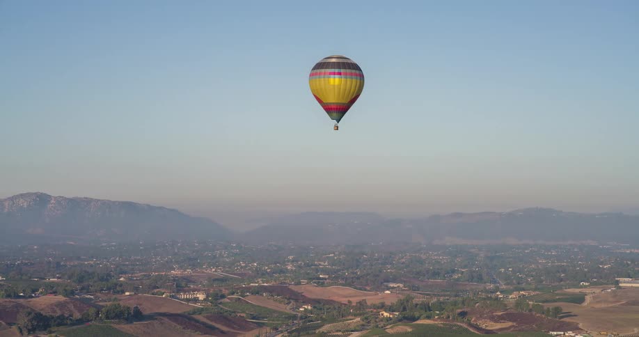 The video shows an aerial shot of a hot air balloon with a yellow and red striped pattern floating over a landscape of rolling hills and vineyards the sun is visible in the background creating a hazy light over the scene